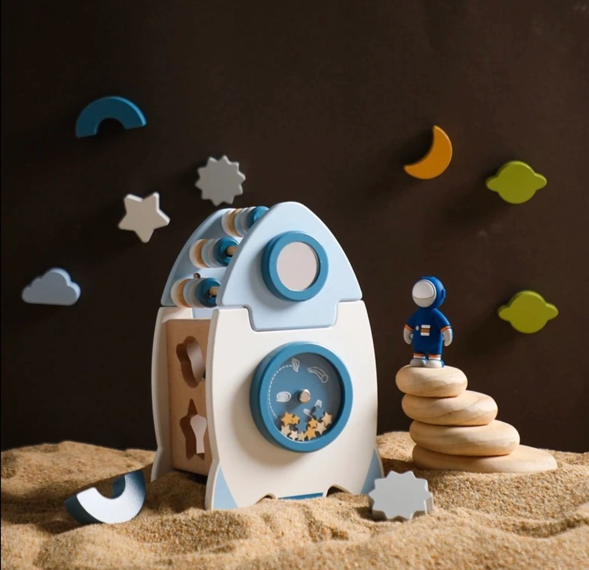 A wooden toy rocket on the sand with a moon and stars in the background.