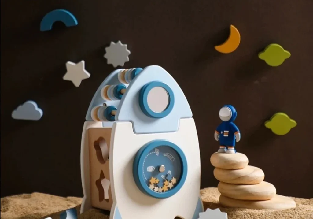 A wooden toy rocket on the sand with a moon and stars in the background.